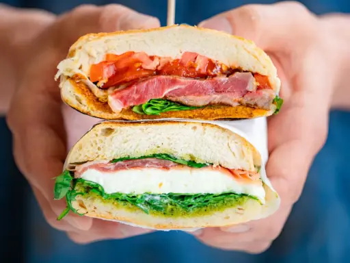 Deux moitiés d'un délicieux sandwich italien, garni de jambon, de tomates, de mozzarella et de pesto, sont tenues par des mains.