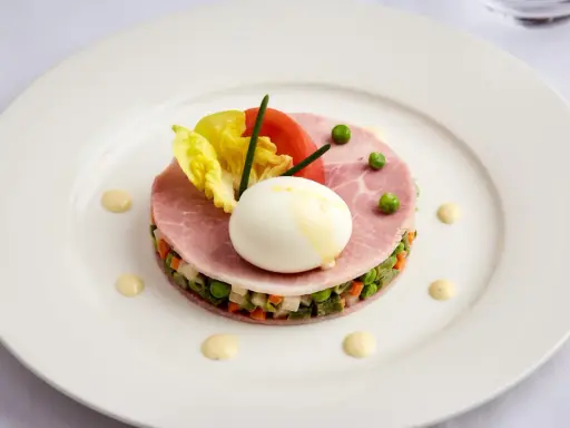 Une assiette blanche présente une délicieuse macédoine de légumes, garnie de jambon, d'un œuf mollet, de tomates et de laitue.