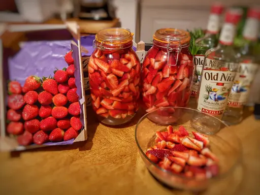 Bocaux de fraises coupées prêtes pour un rhum arrangé fraise menthe. Bouteilles de rhum à proximité.