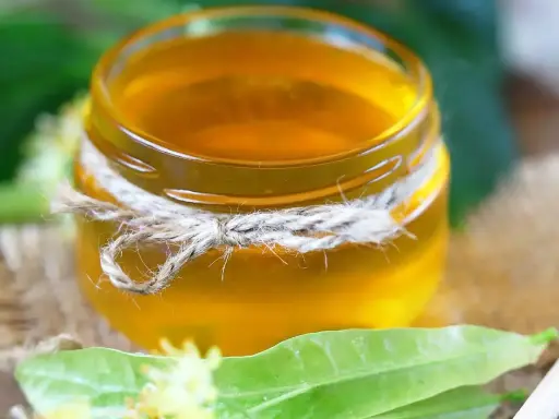 Un pot de sirop de tilleul doré, noué de cordelette, entouré de feuilles vertes et de fleurs de tilleul.