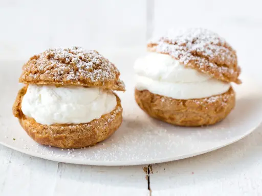 Deux choux à la crème généreusement garnis de crème fouettée, saupoudrés de sucre glace sur une assiette blanche.