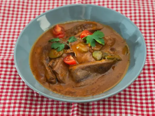 Langue de bœuf en sauce piquante, garnie de cornichons, tomates cerises et persil, servie dans un bol bleu sur une nappe à carreaux rouges et blancs.