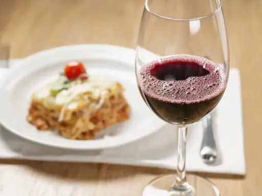 Un verre de vin rouge pétillant à côté d'une portion de lasagnes appétissante. La question se pose : quel vin avec des lasagnes pour un accord parfait ?
