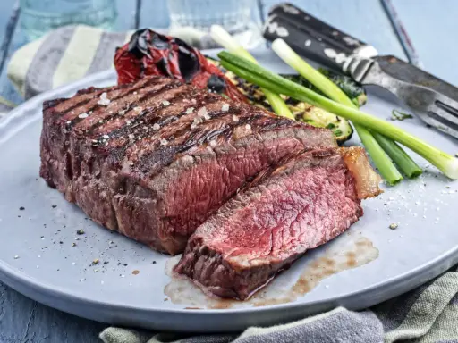 Un savoureux rumsteck de boeuf grillé, coupé en tranches, servi avec des légumes rôtis et des oignons verts sur une assiette grise.