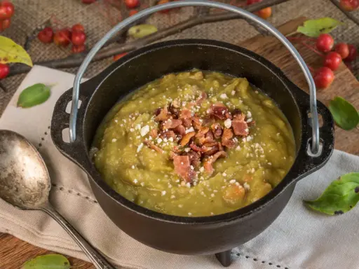 Soupe de pois cassés réconfortante, garnie de lardons croustillants et de fromage râpé, servie dans une cocotte en fonte rustique.