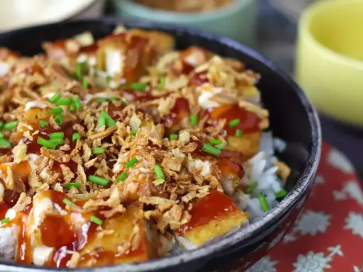 Un bol de riz garni de morceaux de crispy de poulet, nappé de sauce et parsemé d'oignons frits et de ciboule.