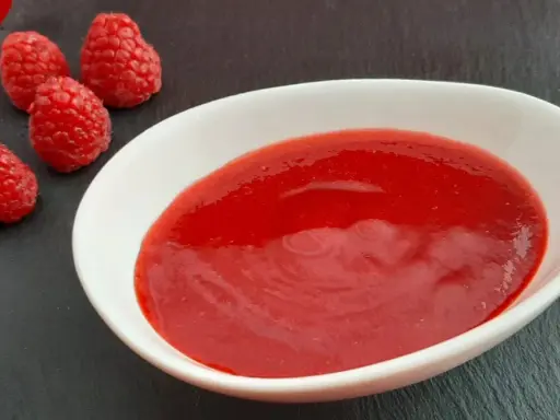 Un bol blanc contient une **purée de framboise** onctueuse, accompagnée de quelques framboises fraîches sur une ardoise.