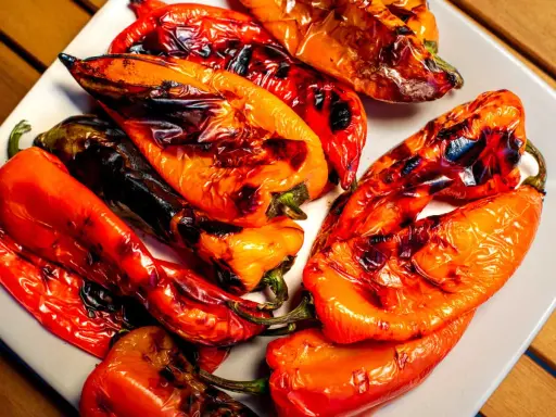 Poivrons rouges et orangés grillés au barbecue, légèrement noircis par la flamme, sur une assiette blanche. Un délice estival.