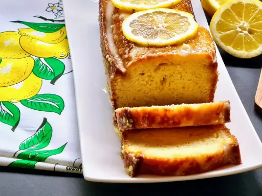 Gâteau moelleux au citron, glacé et garni de rondelles de citron. Ce délice, un gâteau huile d'olive, est servi sur une assiette blanche avec un torchon orné de citrons.