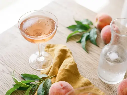 Un verre de vin de pêche rosé, des pêches fraîches et une carafe d'eau glacée sur une table en bois.