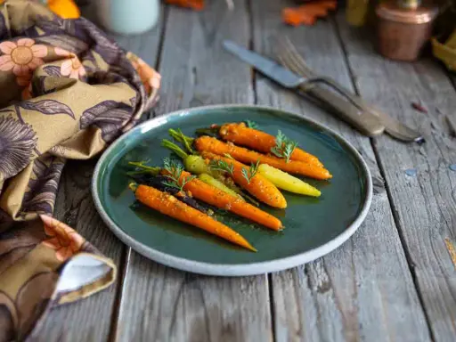 Carottes glacées au miel, colorées et appétissantes, garnissent une assiette sur une table en bois rustique. Une serviette à motifs floraux complète la scène.