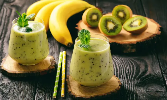 Deux verres de smoothie banane kiwi, décorés de menthe, avec des bananes et des kiwis coupés en arrière-plan.