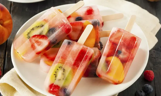 Plusieurs bâtonnets de glace à l'eau fruitée, remplis de fraises, kiwis, pêches et myrtilles, sur une assiette blanche.