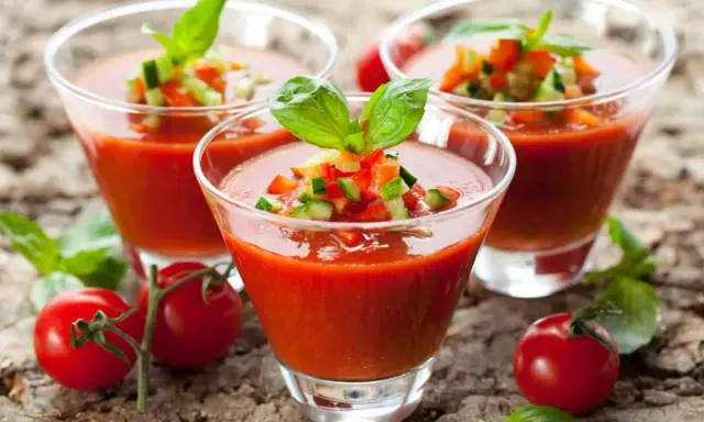 Trois verres de gaspacho tomate, garni de dés de légumes et de basilic, sur fond de bois.