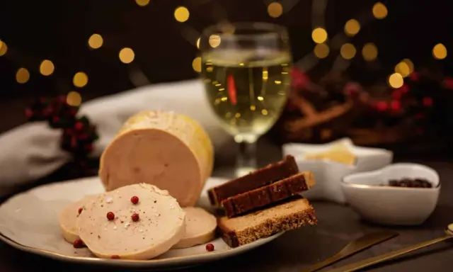 Tranche de foie gras sur une assiette, accompagnée de pain d'épices et d'un verre de vin blanc. Ambiance festive avec des lumières scintillantes.