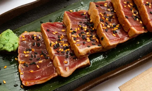 Tranches de tataki de boeuf, poêlé et saupoudré de graines de sésame noires et dorées, servi avec une touche de wasabi sur une feuille verte.