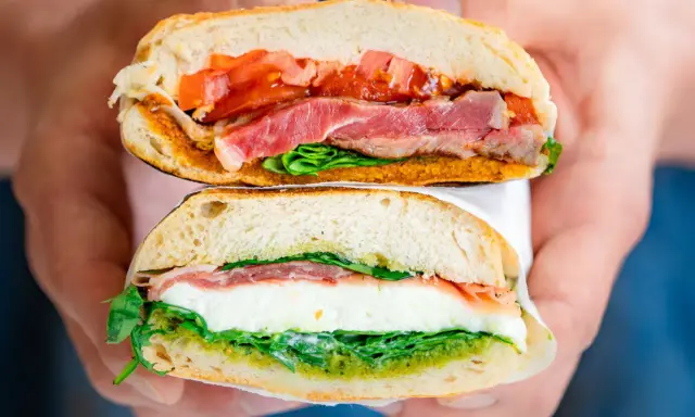 Deux moitiés d'un délicieux sandwich italien, garni de jambon, de tomates, de mozzarella et de pesto, sont tenues par des mains.
