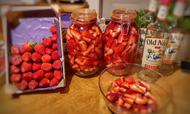 Bocaux de fraises coupées prêtes pour un rhum arrangé fraise menthe. Bouteilles de rhum à proximité.