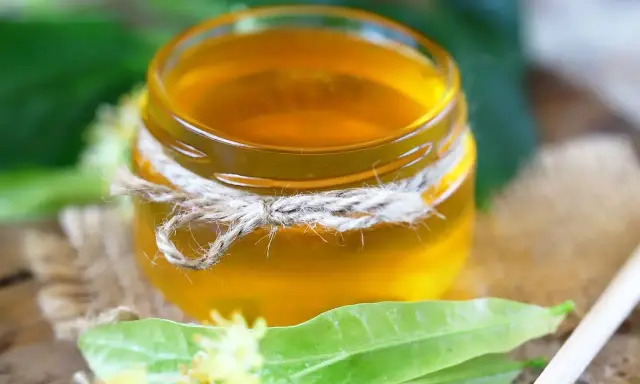 Un pot de sirop de tilleul doré, noué de cordelette, entouré de feuilles vertes et de fleurs de tilleul.