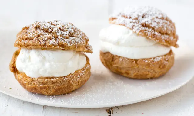 Deux choux à la crème généreusement garnis de crème fouettée, saupoudrés de sucre glace sur une assiette blanche.