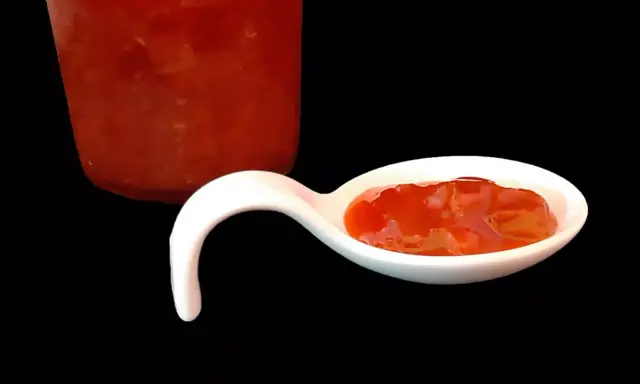Une cuillère blanche présente une délicieuse confiture fraise rhubarbe, avec un pot en arrière-plan.