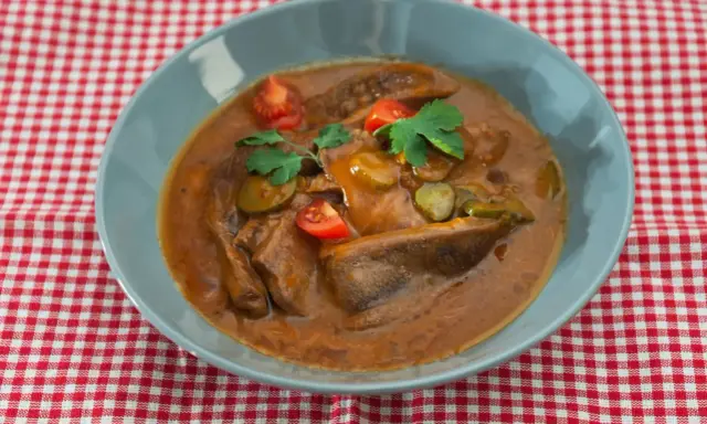 Langue de bœuf en sauce piquante, garnie de cornichons, tomates cerises et persil, servie dans un bol bleu sur une nappe à carreaux rouges et blancs.