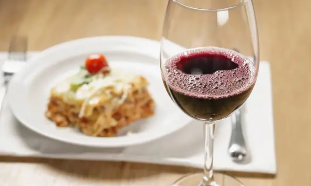 Un verre de vin rouge pétillant à côté d'une portion de lasagnes appétissante. La question se pose : quel vin avec des lasagnes pour un accord parfait ?