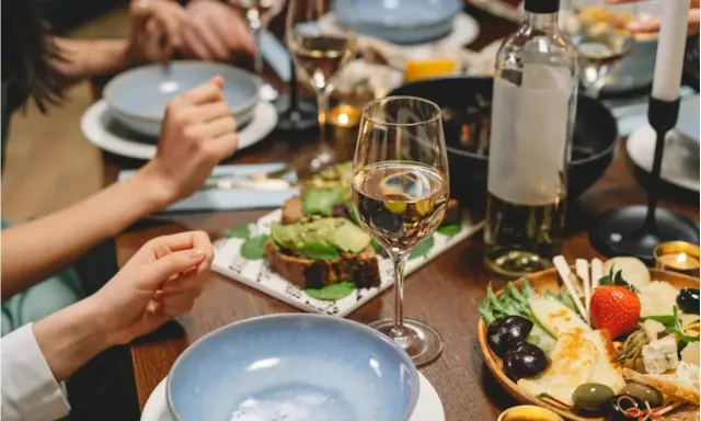 Un verre de vin blanc beurré, des toasts à l'avocat et un plateau de fromages et fruits sur une table dressée pour un dîner convivial.