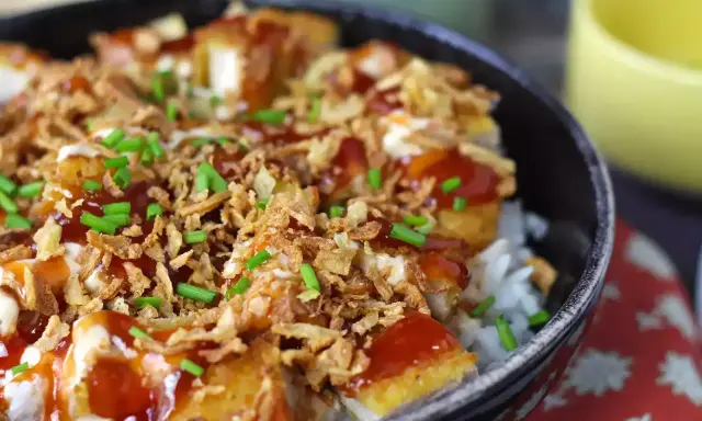 Un bol de riz garni de morceaux de crispy de poulet, nappé de sauce et parsemé d'oignons frits et de ciboule.