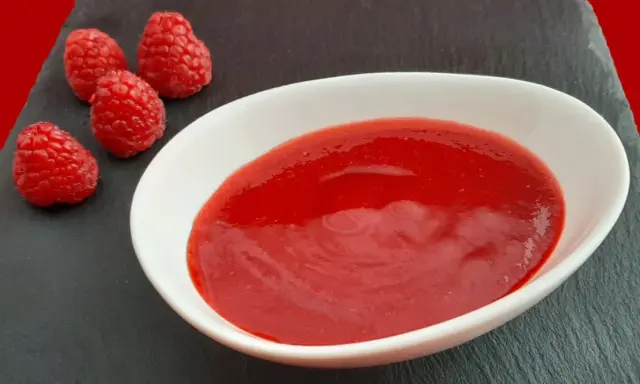 Un bol blanc contient une **purée de framboise** onctueuse, accompagnée de quelques framboises fraîches sur une ardoise.