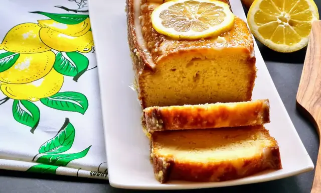 Gâteau moelleux au citron, glacé et garni de rondelles de citron. Ce délice, un gâteau huile d'olive, est servi sur une assiette blanche avec un torchon orné de citrons.