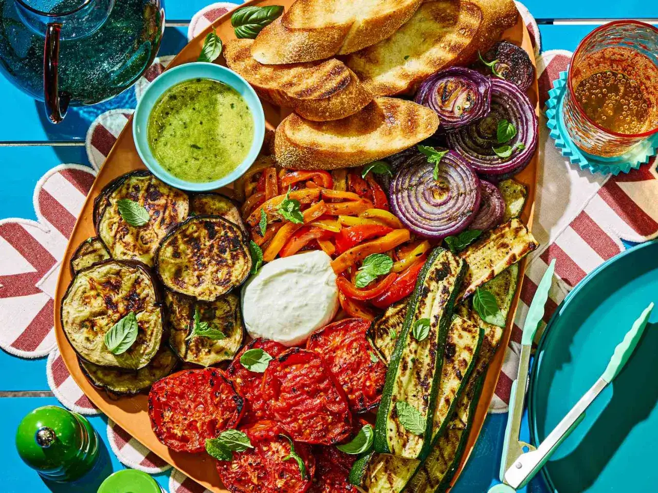 Un plateau coloré de mélange de légumes au four grillés : aubergines, courgettes, poivrons, oignons rouges, tomates, pain grillé et sauce verte.
