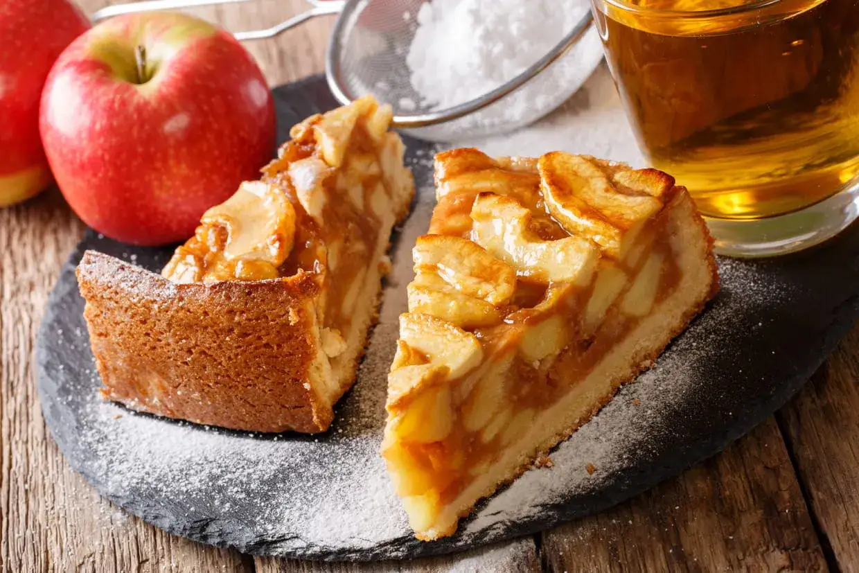 Deux parts de tarte aux pommes, saupoudrées de sucre glace, avec des pommes fraîches et un verre de cidre. Idéal pour savoir que faire avec des pommes !