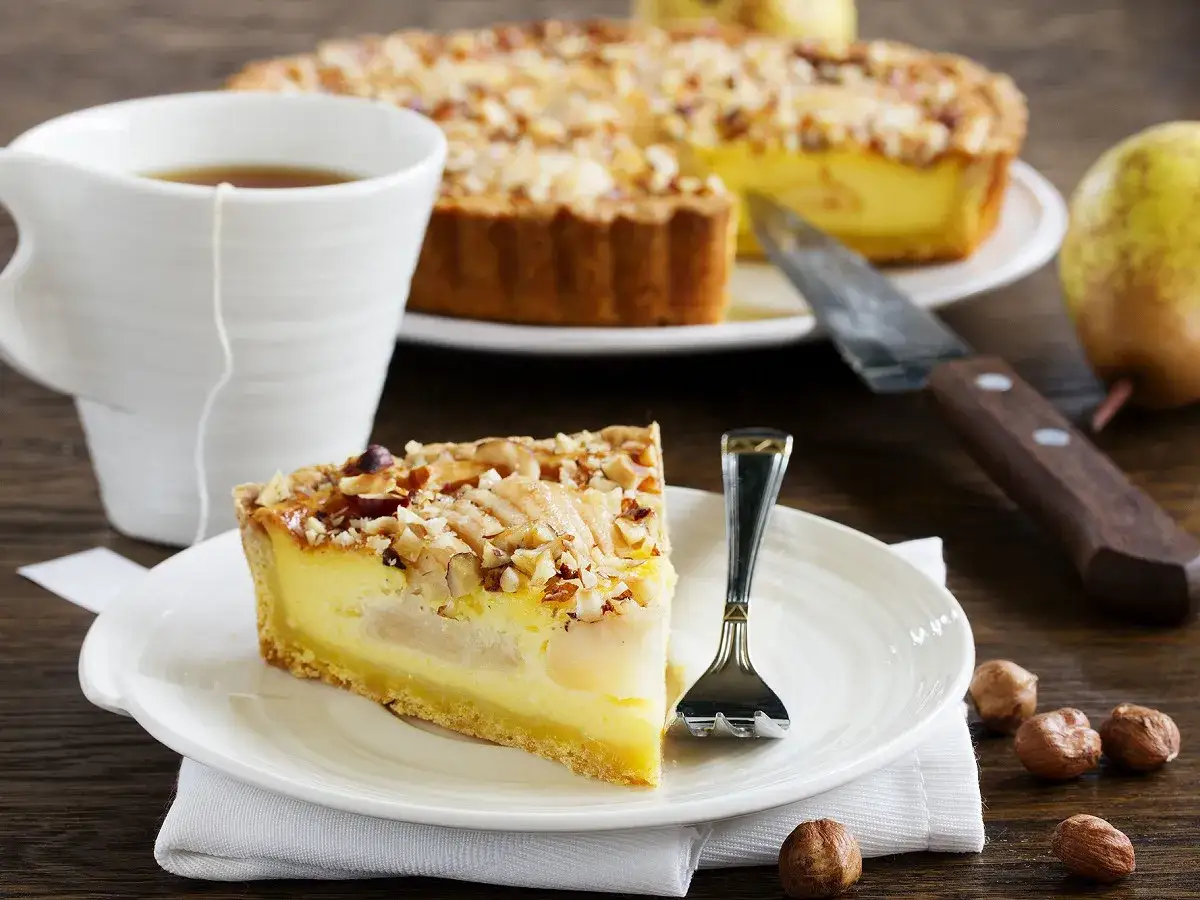 Une part de tarte amandine aux poires, garnie de noisettes concassées, servie avec une tasse de thé et des noisettes entières.