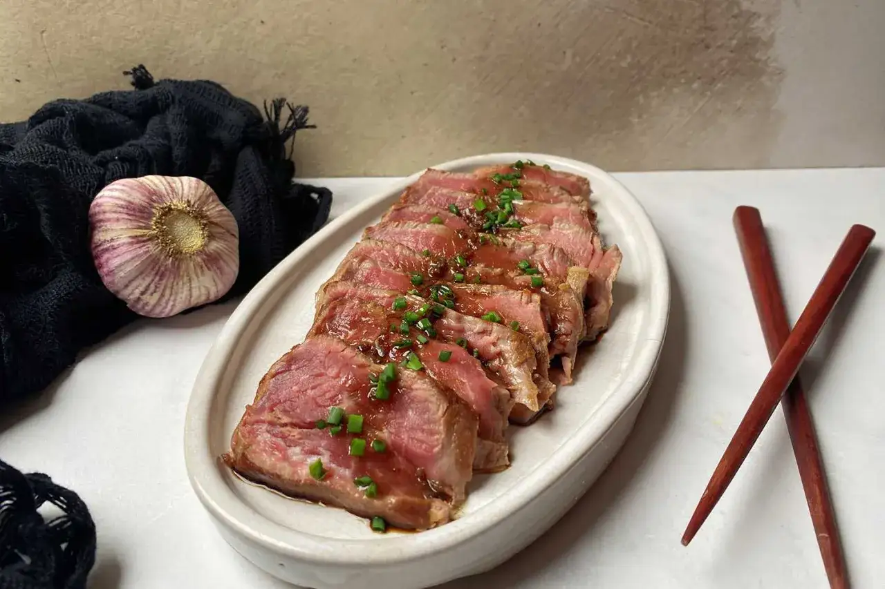 Délicieux tataki de boeuf tranché, nappé d'une sauce savoureuse et garni de ciboule. Une tête d'ail et des baguettes complètent la scène.