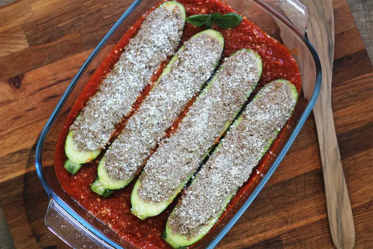 Quatre courgettes farcies à la viande hachée, prêtes à cuire dans une sauce tomate, garnies de parmesan râpé.