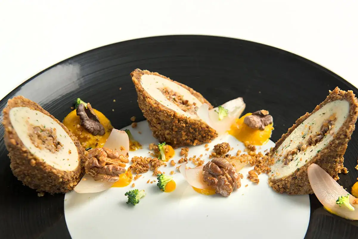 Ballotine de volaille panée, garnie de farce, servie avec purée de courge, noix et brocolis sur une assiette noire.