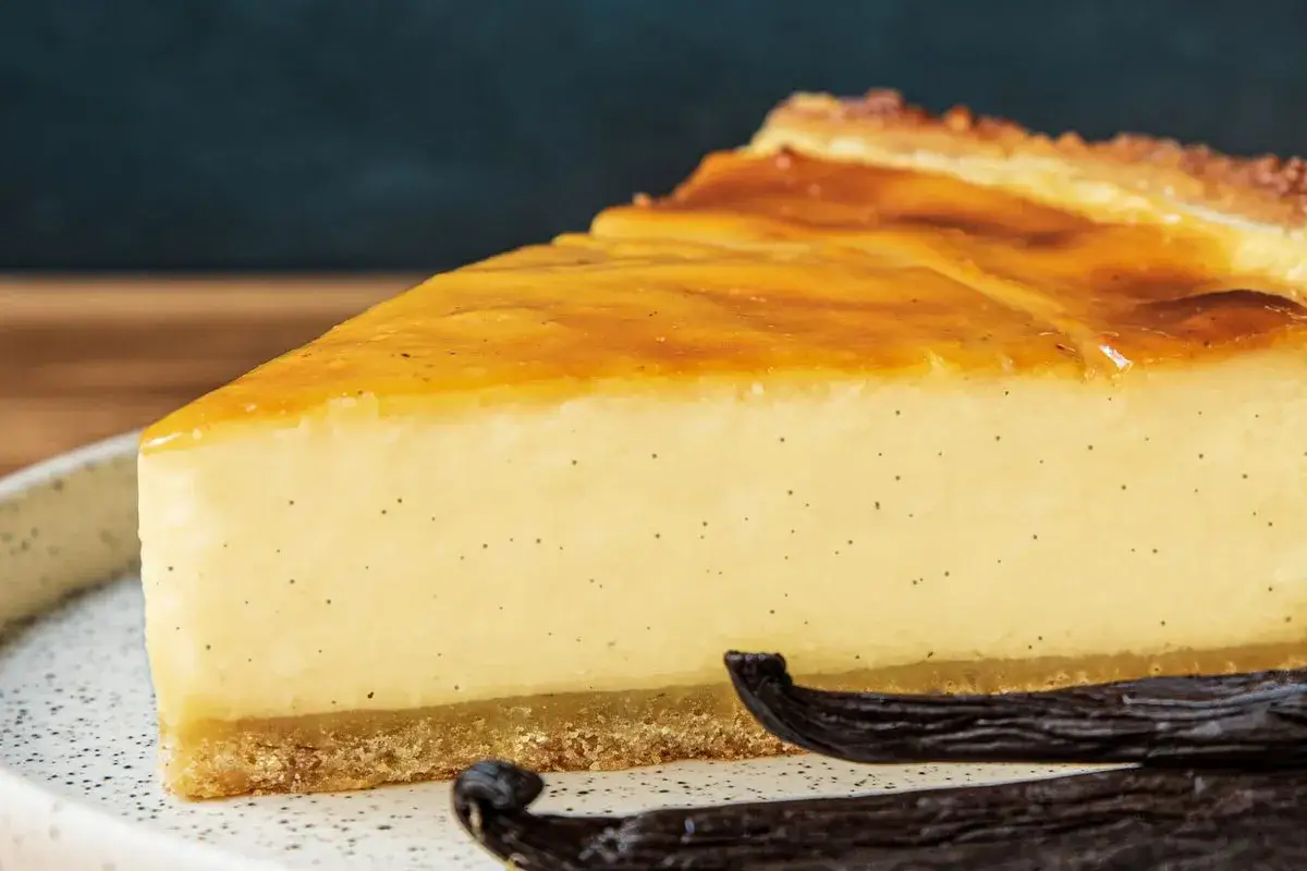 Tranche de flan pâtissier onctueux, parsemé de points de vanille, avec une croûte dorée et des gousses de vanille en premier plan.