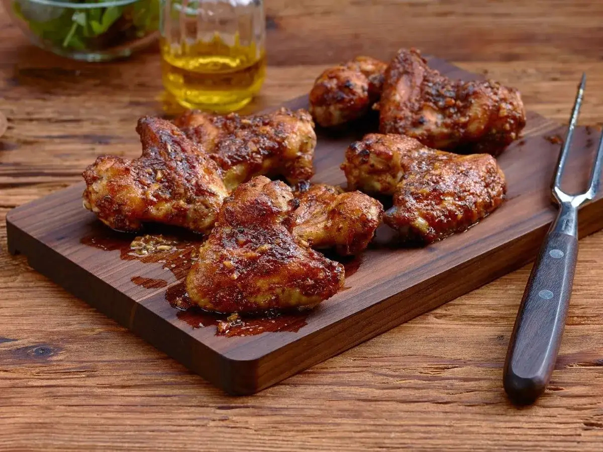 Un plateau de d&eacute;licieux ailerons de poulet grill&eacute;s, brillants de sauce, sur une planche en bois. Une fourchette &agrave; viande est pos&eacute;e &agrave; c&ocirc;t&eacute;.