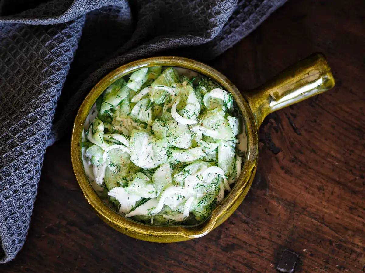Salade fraîche de concombre à la crème, garnie d'aneth et d'oignons, servie dans un bol en céramique sur une table en bois.