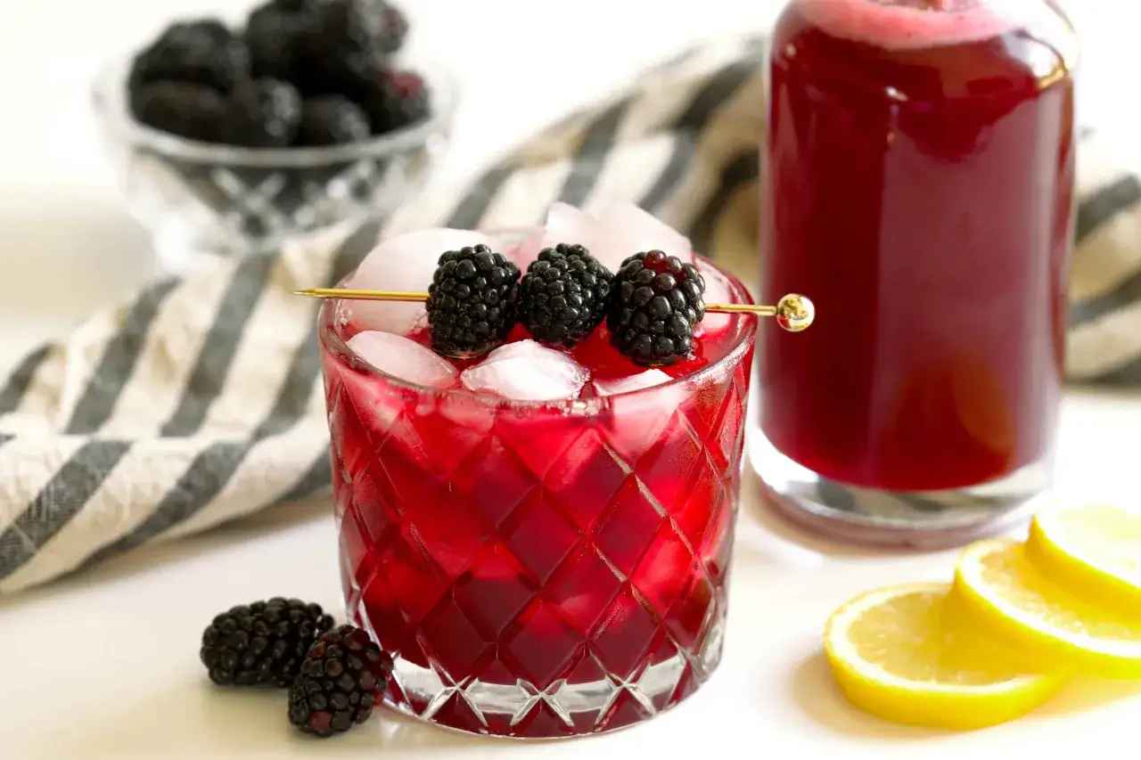 Verre de sirop de mûres glacé, garni de mûres fraîches. Une bouteille de sirop et des rondelles de citron sont visibles en arrière-plan.