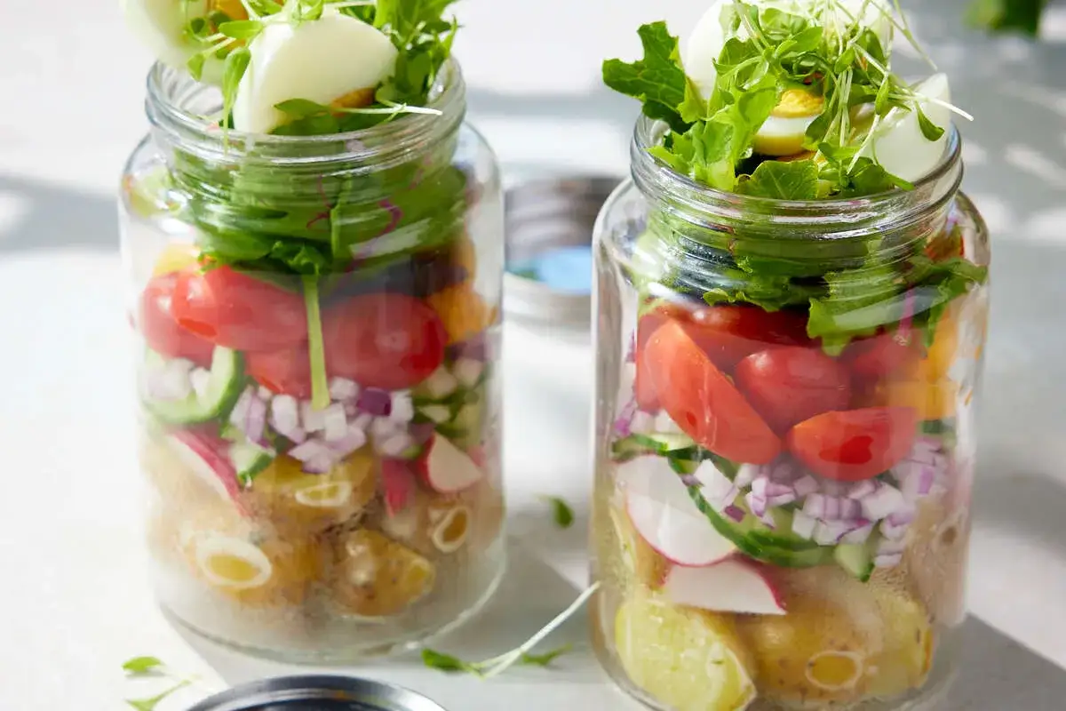 Deux salades pique nique colorées dans des bocaux en verre, garnies de pommes de terre, tomates, concombres, radis, oignons rouges, œufs durs et jeunes pousses.