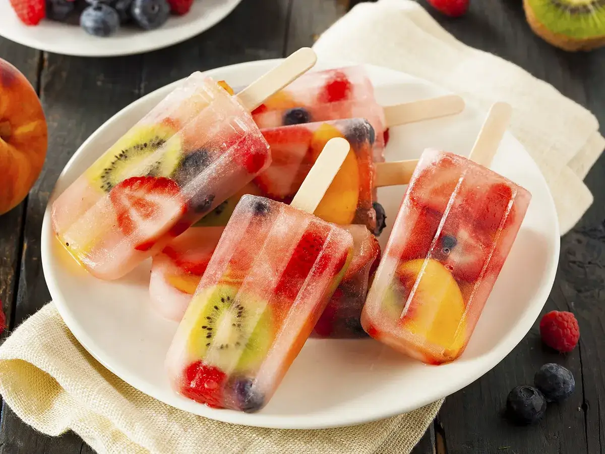 Plusieurs bâtonnets de glace à l'eau aux fruits, avec des fraises, des kiwis, des pêches et des myrtilles, sur une assiette blanche.