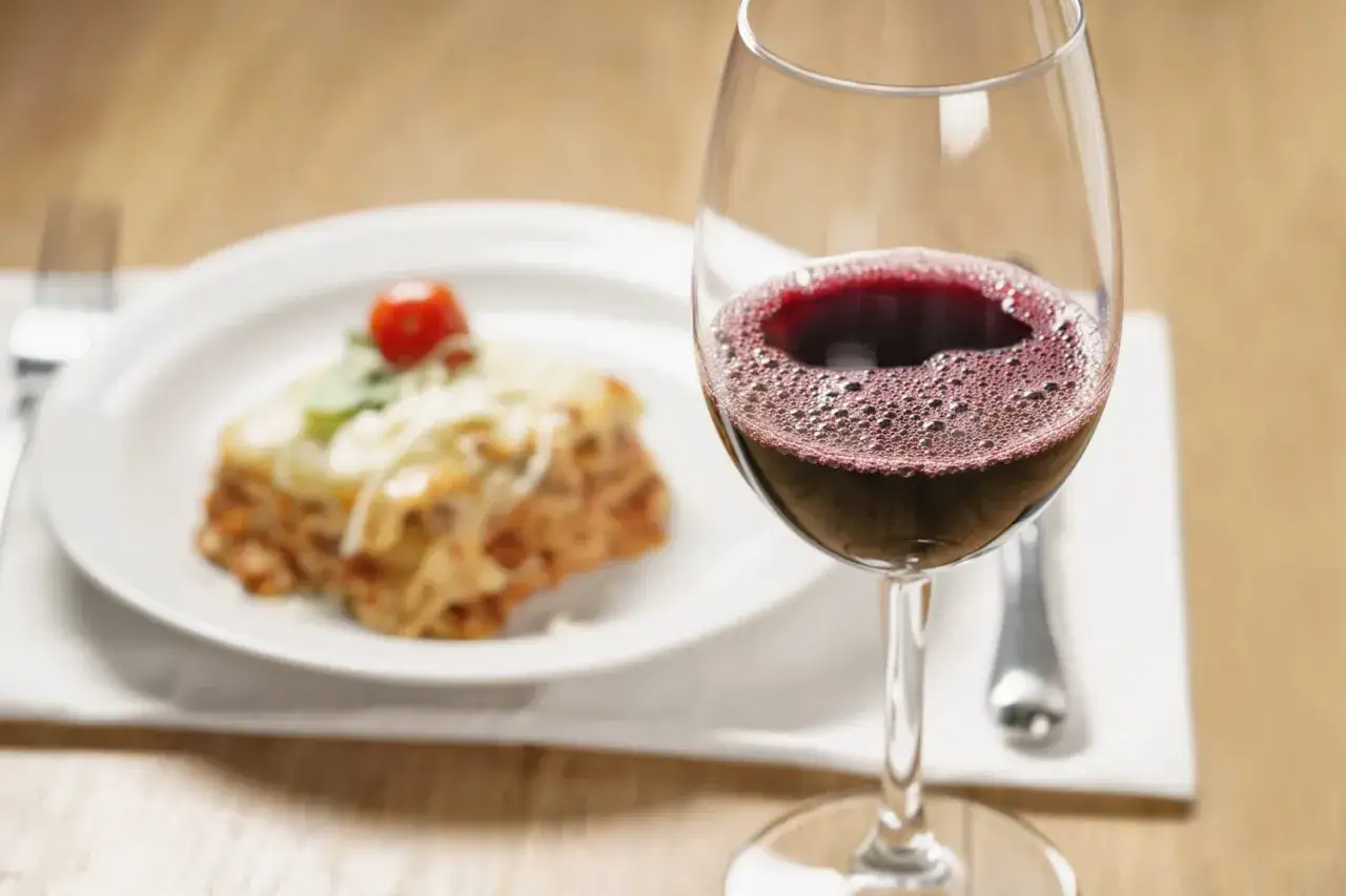 Un verre de vin rouge pétillant à côté d'une lasagne appétissante. La question est : quel vin avec des lasagnes pour un repas parfait ?