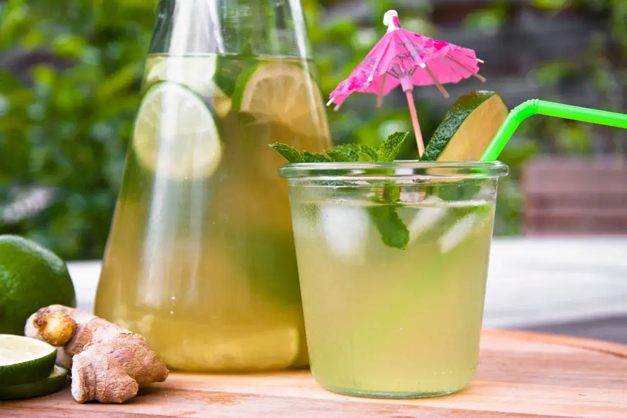 Un verre de mocktail ginger beer rafraîchissant, garni de menthe et d'une tranche de citron vert, avec une carafe remplie de la même boisson en arrière-plan.