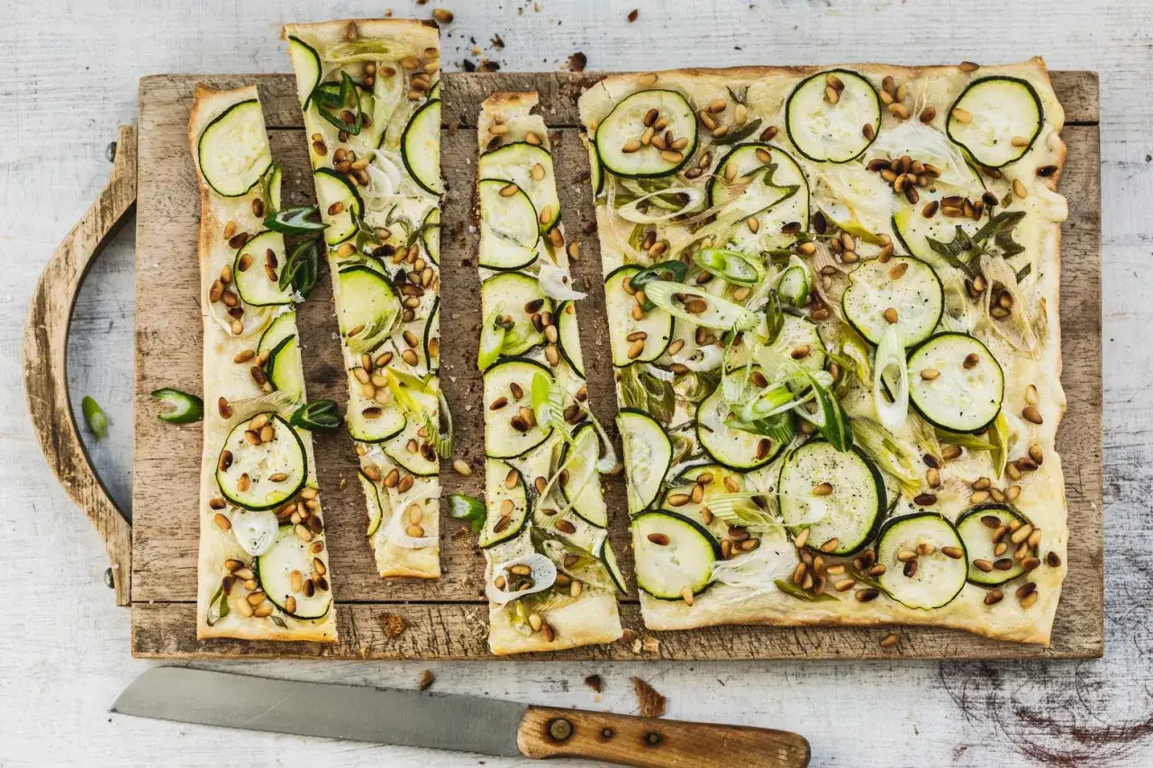 Délicieuse tarte flambée végétarienne aux courgettes, oignons nouveaux et pignons de pin, coupée en fines lamelles sur une planche en bois.