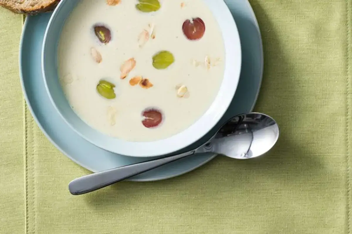 Un bol de ajo blanco crémeux garni de raisins verts et rouges, d'amandes effilées et de quelques éclats de noisettes. Une cuillère repose à côté.