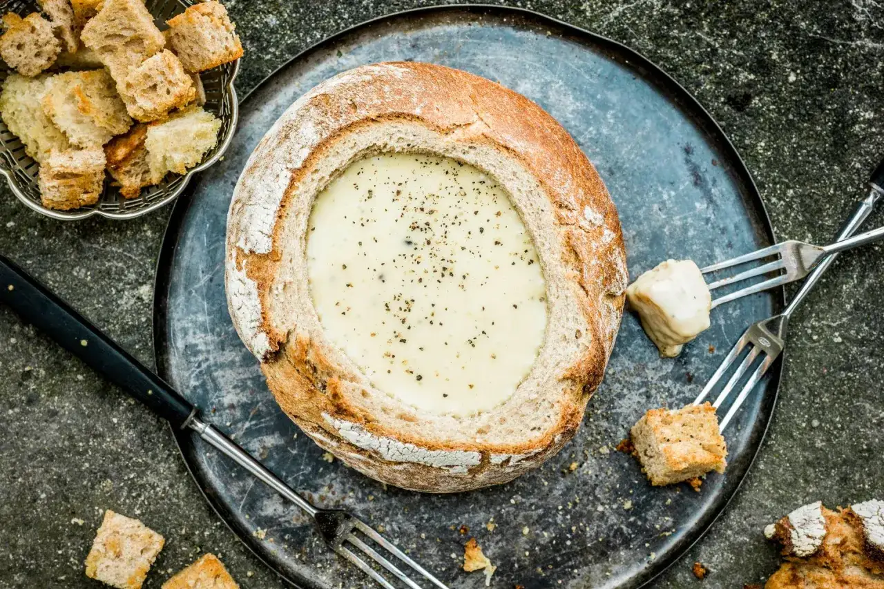 Fondue savoyarde cr&eacute;meuse dans un pain rond, servie avec des cro&ucirc;tons et du vin blanc.