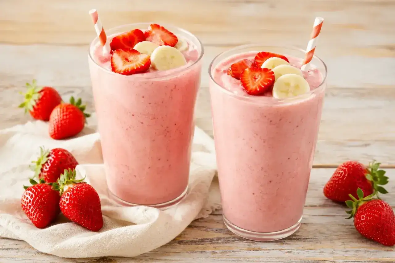 Deux verres de smoothie fraise banane, garnis de fruits frais et de pailles rayées, sur une table en bois.