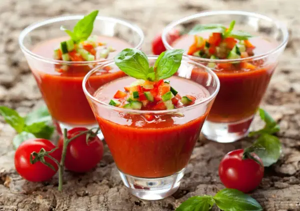Trois verres de gaspacho tomate, garni de dés de légumes et de basilic, sur fond de bois.